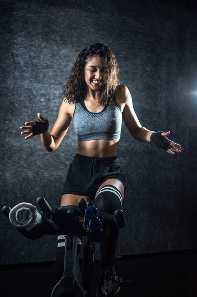 Cheerful sporty woman dancing on exercise bike during cycling ride indoors. - Photo, Image