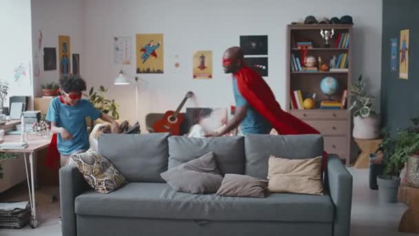 Медленная съемка возбужденного афроамериканского отца в накидке супергероя и маске, гоняющегося за маленьким сыном, бегающим по гостиной, играя вместе дома - Кадры, видео