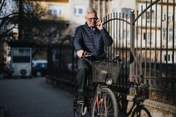 Aktivní senior profesionál v bundě bilance na kole, zatímco zapojený do hovoru na svém mobilním telefonu, symbolizující multitasking a ekologicky šetrné dopravy ve městě. - Fotografie, Obrázek