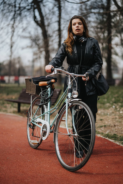 Una mujer de negocios confiada con auriculares camina con su bicicleta en un parque de la ciudad, encarnando desplazamientos urbanos respetuosos con el medio ambiente. - Foto, imagen