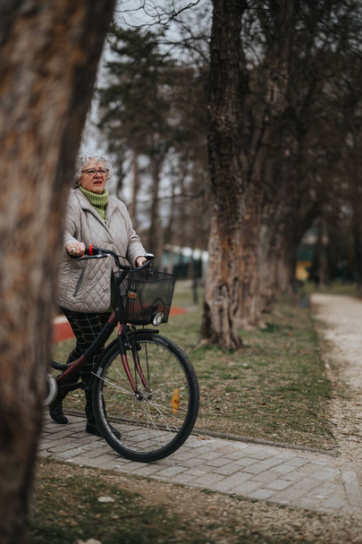 Mature female retiree with glasses and casual attire takes a break from cycling in a serene park setting. - Photo, Image