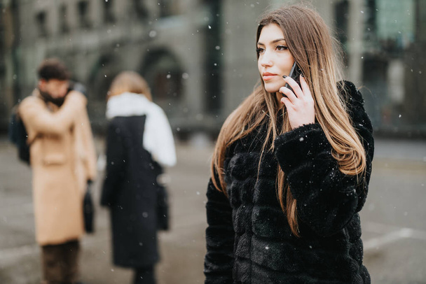 Mujer joven enfocada hablando por teléfono, con colegas borrosos en el fondo en una calle urbana nevada. - Foto, Imagen