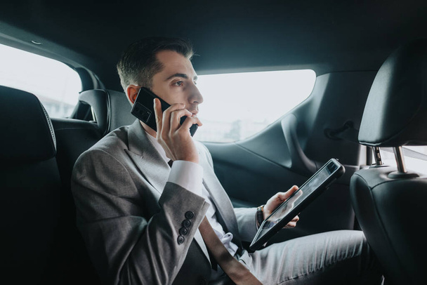 Un profesional de negocios bien vestido sentado en el asiento trasero de un vehículo, posiblemente de camino a una reunión. - Foto, imagen