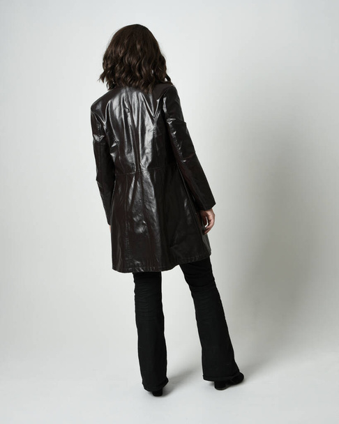 Full length portrait of brunette woman wearing long leather trench coat and black boots. Standing pose walking away from camera, facing backwards. Isolated silhouette on white studio background. - Photo, Image