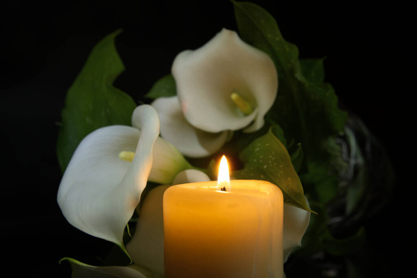 Flor de lirio de cala blanca junto a la llama de la vela brillante arroja una luz cálida en un ambiente oscuro y crea un ambiente relajante, irradiando una sensación de soledad pacífica - Foto, Imagen