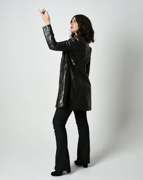Full length portrait of brunette woman wearing long leather trench coat. Standing pose walking away from camera, looking back over shoulder, reaching out. Isolated on white studio background. - Photo, Image