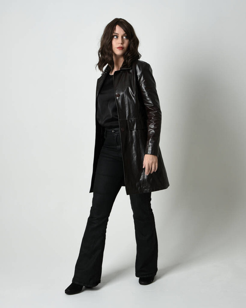 Full length portrait of beautiful brunette woman wearing long black leather trench coat, film noir detective. Standing pose, walking towards the camera, reaching out. Isolated white studio background - Photo, Image