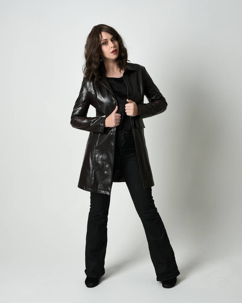 Full length portrait of beautiful brunette woman wearing long black leather trench coat, film noir detective. Standing pose, walking towards the camera, reaching out. Isolated white studio background - Φωτογραφία, εικόνα
