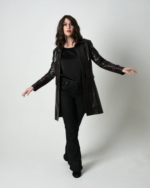 Full length portrait of beautiful brunette woman wearing long black leather trench coat, film noir detective. Standing pose, walking towards the camera, reaching out. Isolated white studio background - Foto, Bild