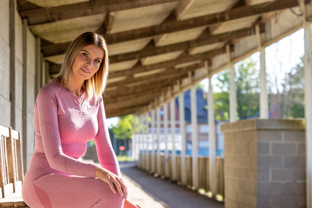 Una atlética en ropa deportiva rosa encuentra un momento de descanso tranquilo en un banco de madera bajo el dosel lineal de una instalación deportiva urbana. Descanso atlético: Mujer en ropa deportiva rosa disfrutando de la soledad urbana - Foto, imagen