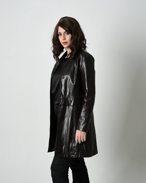 close up portrait of brunette woman wearing long leather trench coat. Standing pose in side profile, looking back over shoulder, reaching out. Isolated on white studio background. - Φωτογραφία, εικόνα