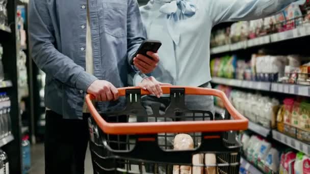 Deux personnes caucasiennes vêtues de vêtements décontractés faisant leurs courses dans une épicerie. Jeune mari tenant smartphone moderne tout en conduisant panier entre les rangées dans le supermarché. Concept d'achat et de famille. - Séquence, vidéo