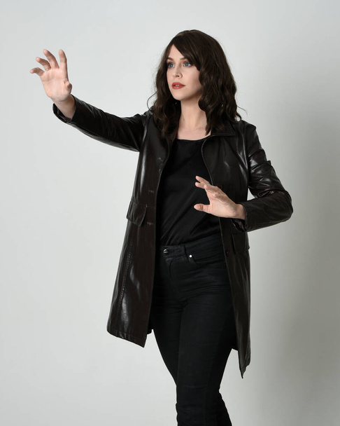 close up portrait of beautiful brunette woman wearing long black leather trench coat, film noir detective.  Gestural hand poses, arms reaching out, Isolated on studio background - Zdjęcie, obraz