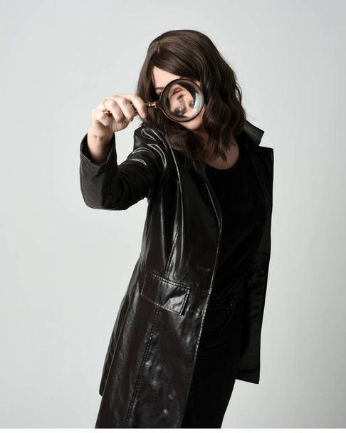 close-up portret van mooie brunette vrouw dragen zwart lederen trench jas. Rechercheur vergrootglas vasthouden, naar de camera reiken, op zoek naar ontdekking. Geïsoleerde studio achtergrond - Foto, afbeelding