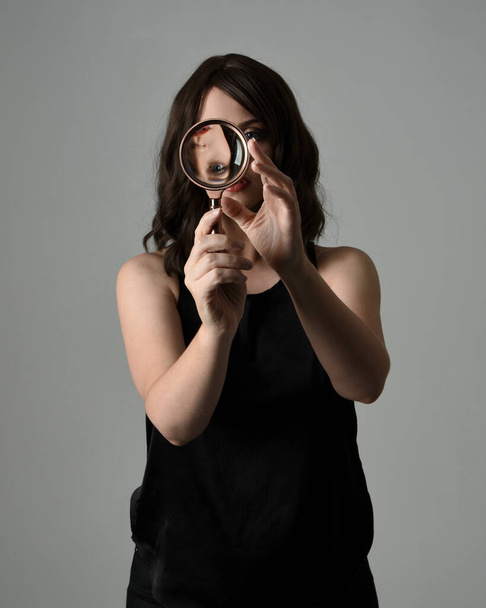 close up portrait of beautiful brunette woman wearing black leather trench coat. Holding detective magnifying glass, reaching out towards camera, searching for discovery. Isolated  studio background - Photo, Image