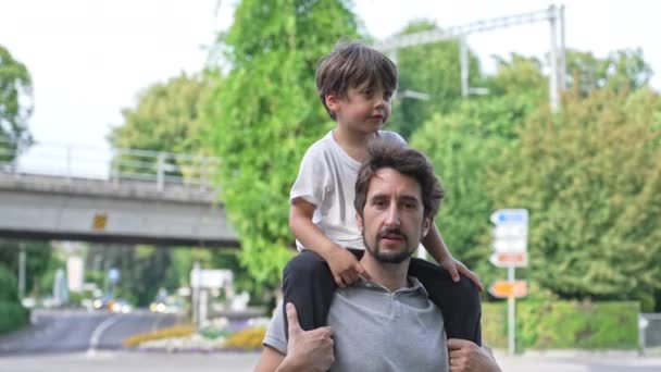 Γιος στον ώμο του πατέρα - Piggyback. Πορτρέτο του γονέα και του παιδιού κοιτάζοντας κάμερα στέκεται έξω σε ηλιόλουστη μέρα στο Sidewalk - Πλάνα, βίντεο