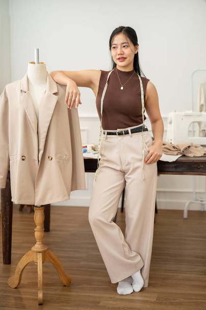 A confident and attractive Asian female fashion designer is standing in her atelier studio with a tailor mannequin or tailor dummy, looking at the camera. - Фото, изображение