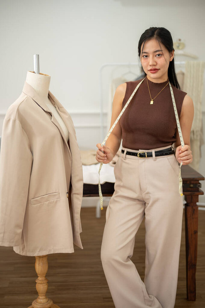 An attractive and confident Asian female tailor or fashion designer is in her studio, standing beside a tailor dummy and looking at the camera. atelier, seamstress, dressmaker, fashion industry - Photo, Image
