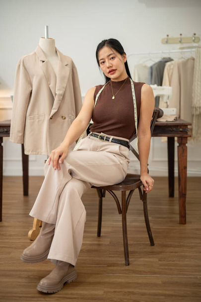 A confident and attractive Asian female fashion designer or tailor is sitting on a chair in her studio, looking at the camera. people and fashion concepts - Photo, Image