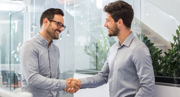 Двоє усміхнених бізнесменів трясуться разом, стоячи в сучасному офісі. - Фото, зображення