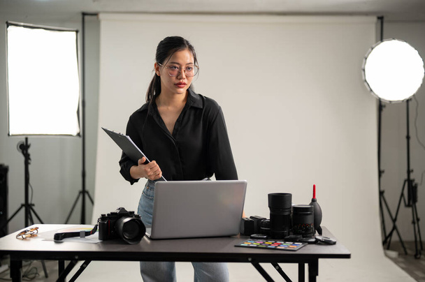 Egy vonzó, elegáns ázsiai női fotós fekete ingben áll a fotós stúdiójában professzionális fényképészeti felszereléssel. - Fotó, kép