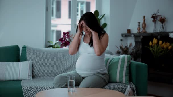 30s kobieta w ciąży zmaga się z bólem głowy i ból siedzi w domu kanapa rezydencja, macierzyńskie koncepcji zdrowia - Materiał filmowy, wideo