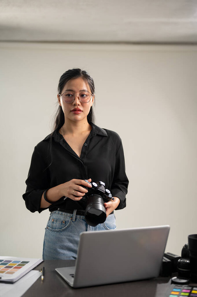 An elegant, professional Asian female photographer in a black shirt with her DSLR camera is standing in her studio. - Photo, Image