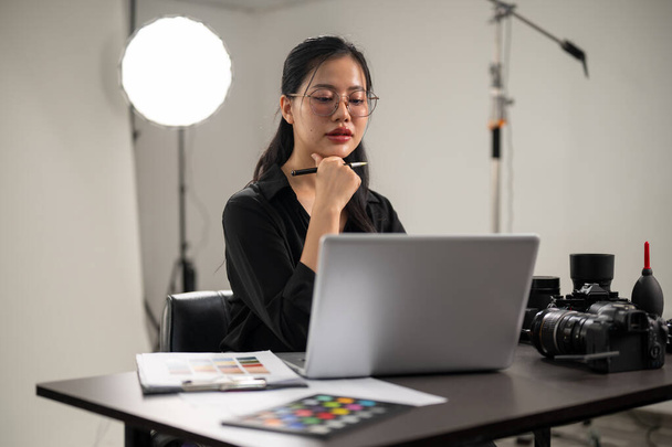 Egy magabiztos, profi ázsiai női fotós dolgozik a laptopján, képeket ellenőriz, egy professzionális világítási berendezéssel rendelkező fotós stúdióban dolgozik.. - Fotó, kép
