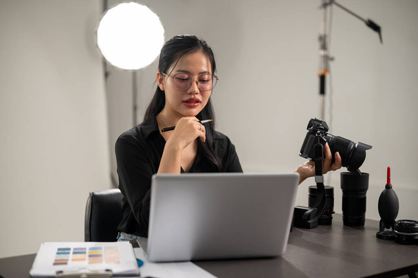Μια επαγγελματίας, κομψή Ασιάτισσα φωτογράφος με μαύρο πουκάμισο εστιάζει στον έλεγχο εικόνων στον φορητό υπολογιστή της, δουλεύοντας σε στούντιο.. - Φωτογραφία, εικόνα