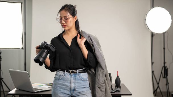 A confident, professional Asian female photographer in a black shirt is standing in her photoshoot studio with a DSLR camera in her hand. - Photo, Image