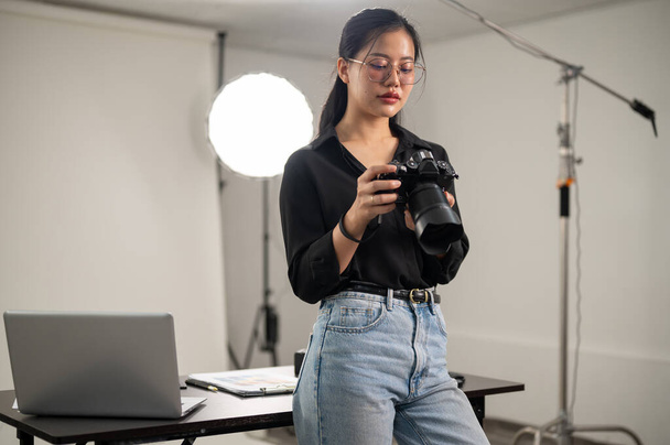 Profesjonalna azjatycka fotografka skupia się na regulacji lustrzanki, pracując w studiu sesyjnym ze sprzętem oświetleniowym. - Zdjęcie, obraz