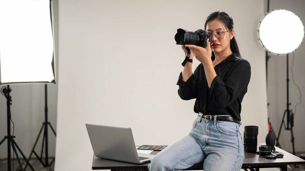 Profesyonel, kendine güvenen bir Asyalı kadın fotoğrafçı fotoğraf çekiyor, yüzünün yanında bir kamera tutuyor, kamerasını ayarlıyor, profesyonel ışıklandırma ekipmanlarıyla bir stüdyoda çalışıyor.. - Fotoğraf, Görsel