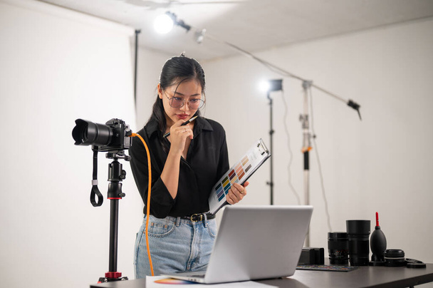 Una atractiva productora de fotógrafos asiática está trabajando en un estudio de fotografía, centrándose en comprobar las imágenes en su computadora portátil. un estudio con equipo profesional de iluminación fotográfica - Foto, Imagen