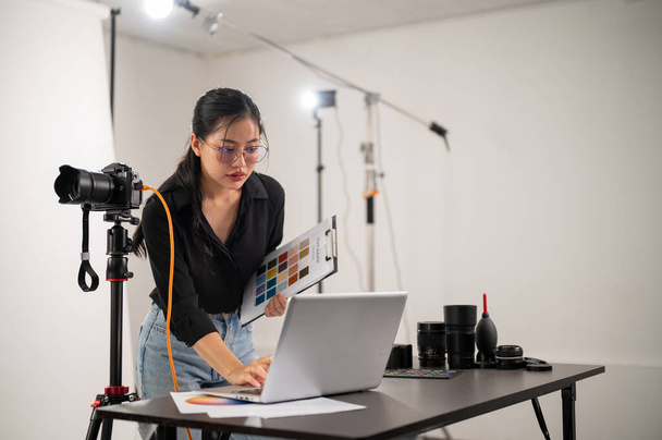 Uma produtora asiática de fotógrafos está trabalhando em um estúdio de tiro, focando em verificar imagens em seu computador portátil. um estúdio com equipamento de iluminação de fotografia profissional - Foto, Imagem