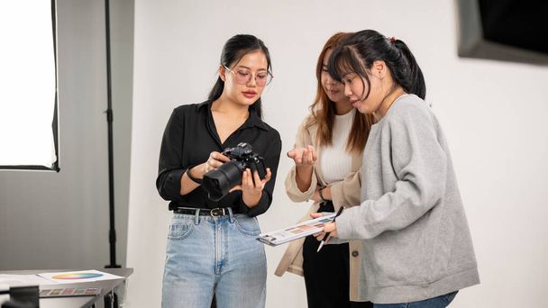 Una fotógrafa asiática profesional y genial está hablando y mostrando imágenes en la cámara a su equipo, trabajando en el estudio de fotografía de moda e informando sobre el concepto con un director de arte.. - Foto, imagen