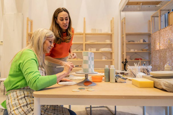 Μια εστιασμένη συνεδρία δύο γυναικών που ζωγραφίζουν γεωμετρικά μοτίβα σε ένα κεραμικό αγγείο σε εργαστήριο χειροτεχνίας. - Φωτογραφία, εικόνα