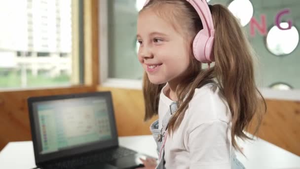 Hymyilevä tyttö etsii samalla heiluttaen käsi kameralle kannettavan sijoitettu pöydälle. Lapsi yllään kuulokkeet hymyilevä kun kannettava näyttö näyttää järjestelmän ohjelmointi tai koodaus ohjelma STEM luokassa. Virheellisyys. - Materiaali, video