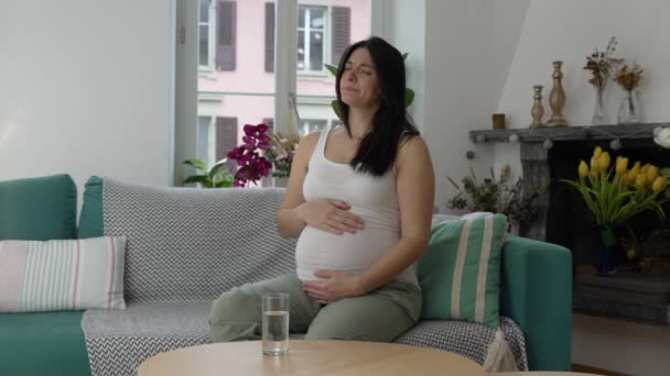 Kobieta w ciąży z bólem, kobieta po trzydziestce walcząca z nudnościami podczas trzeciego trymestru ciąży, siedząca na kanapie w domu, delikatnie dotykająca brzucha. - Materiał filmowy, wideo