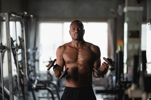 ジムでダンベル運動をしているアフリカのスポーツマン。筋肉トレーニングのためのアジアの体の建物。スポーツ健康ジムのコンセプト - 写真・画像