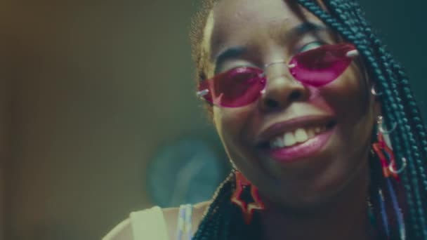 Slowmo medium zbliżenie portret stylowa Afroamerykanka młoda kobieta w pleciony styl życia i różowe okulary pozowanie do kamery i taniec w latarce wewnątrz - Materiał filmowy, wideo