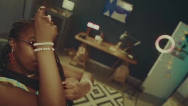 Foto de mano de la chica afroamericana de lujo con peinado trenzado y traje y2k mirando a la cámara a través de gafas de color rosa vintage y bailando en el interior de un apartamento de luz cálida - Metraje, vídeo