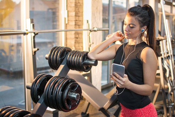 Спортивная девушка с наушниками, слушающая музыку на смартфоне в спортзале. - Фото, изображение