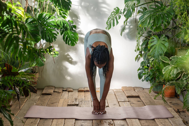 Перед фитнес-тренировкой, стоя на коврике для йоги во внутренних городских джунглях, стройная африканская девушка делает наклоны тела вперед для гибкости. Предварительная подготовка разминка, здоровый образ жизни. - Фото, изображение