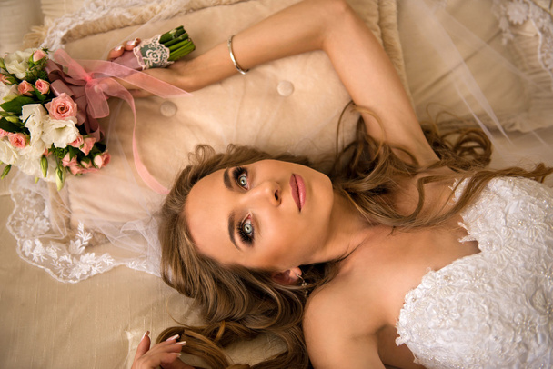 Charming beautiful bride holding wedding bouquet in her hands. bride in white dress in a luxury interior on her back on the beautiful white bed. beautiful bride in a long white veil - Fotografie, Obrázek