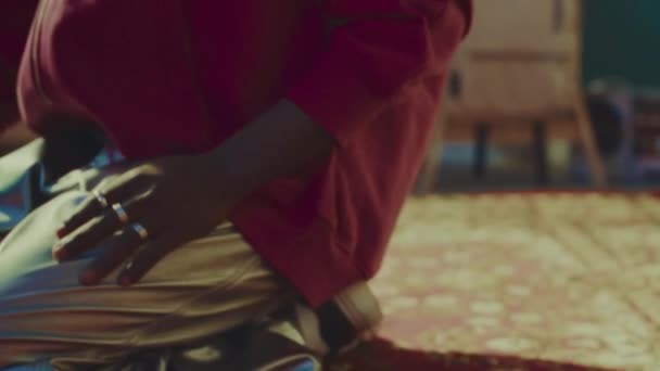 Portrait incliné de gen z Noir en pantalon argenté, chapeau seau en fourrure rose et lunettes de soleil jaunes posant pour appareil photo dans un appartement de style vintage avec tapis rouge sur le sol - Séquence, vidéo