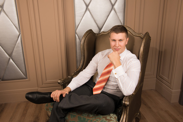 Vertrouwen knappe zakenman in chique interieur. Succesvolle jonge man in een pak zitten op de stoel met zijn benen gekruist en glimlachen - Foto, afbeelding