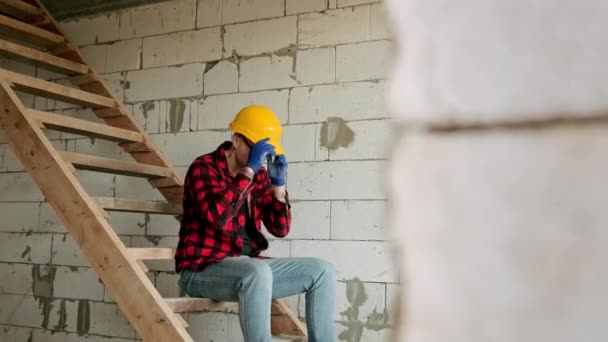 Trabajador de la construcción cansado sentado en las escaleras contra la pared de ladrillo quitándose el sombrero duro y gafas mientras mira a su alrededor. Movimiento lento - Imágenes, Vídeo