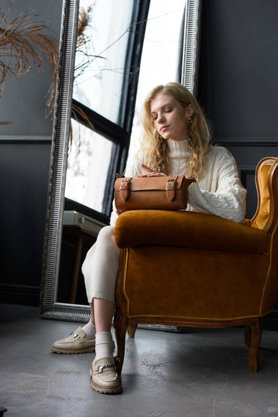 schöne Frau mit blonden Locken posiert mit einer kleinen braunen Tasche in einem Vintage-Stuhl. Model trägt stylischen weißen Pullover und klassische Hose - Foto, Bild