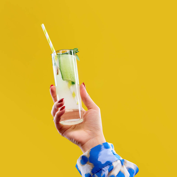 Женщина держит стакан джин-тоника с огурцом и полосатой соломой на ярком желтом фоне студии. Концепция алкоголя, напитков, вечеринок, дегустаций, праздников. Копирование места для рекламы - Фото, изображение
