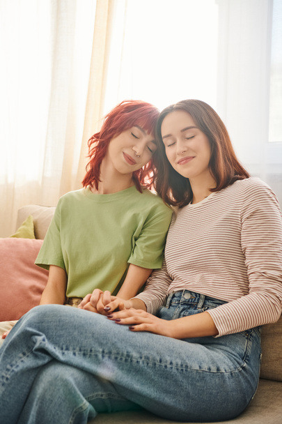 tenderness, young and pleased lesbian couple in casual attire holding hands and sitting on sofa - Photo, Image